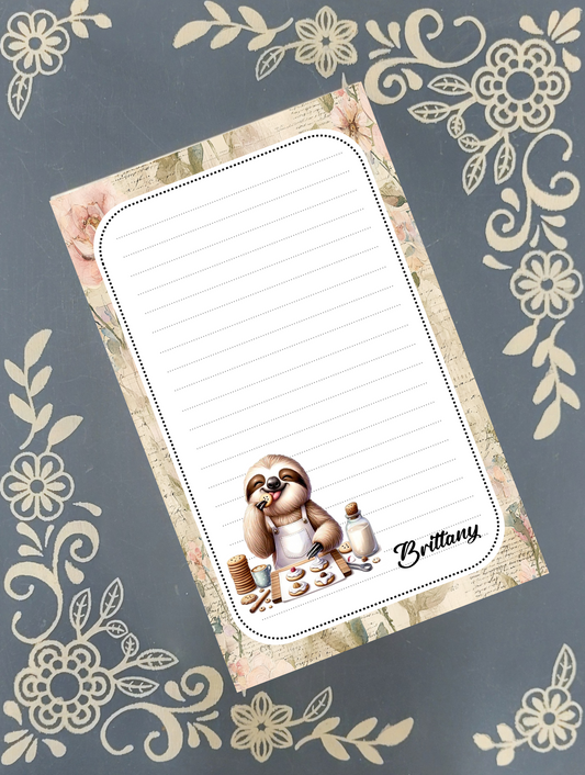 Personalized notepad featuring a whimsical illustration of a baking sloth wearing a chef hat holding a cookie with custom name text , placed on a dark gray background with cream floral patterns. The name 'Brittany' appears in script at the lower right of the notepad.