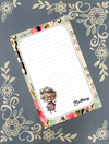 A personalized lined notepad with a floral and musical patterned border, featuring a cartoon illustration of an elderly woman and the name 'Brittany' in script. The notepad is displayed diagonally on a dark gray background decorated with cream-colored floral motifs.