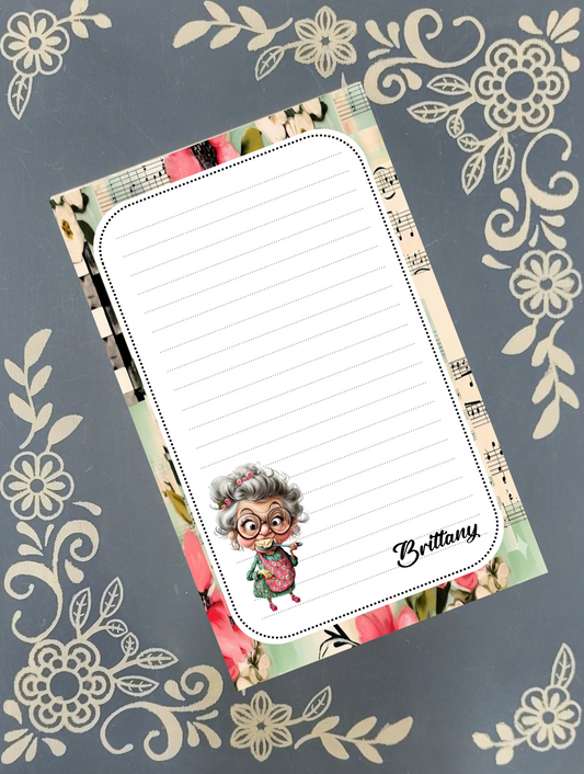 A personalized lined notepad with a floral and musical patterned border, featuring a cartoon illustration of an elderly woman and the name 'Brittany' in script. The notepad is displayed diagonally on a dark gray background decorated with cream-colored floral motifs.