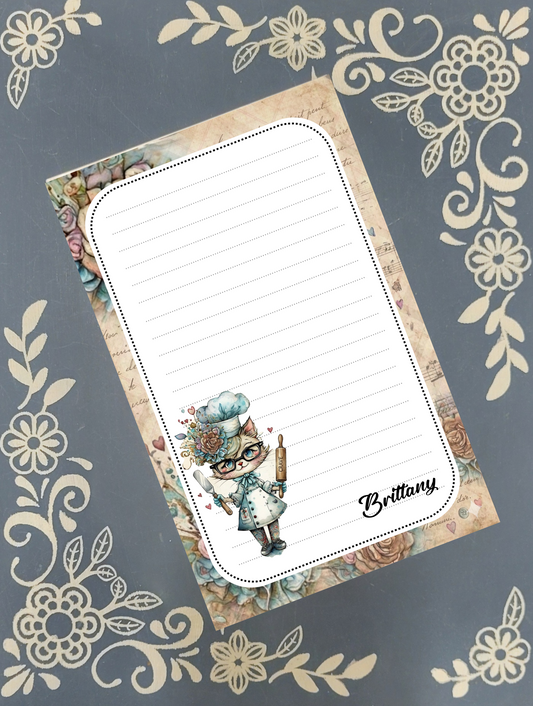 A rectangular lined notepad with a vintage floral border and an illustration of a cat wearing a chef's hat and holding a rolling pin, placed on a gray background with cream floral patterns. The name 'Brittany' is printed in script near the bottom right.
