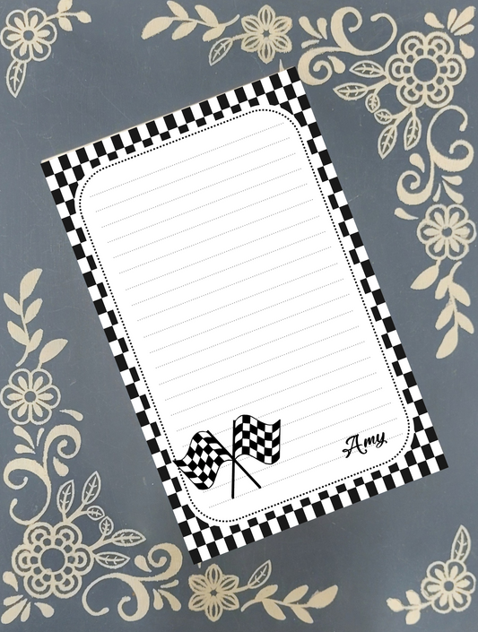 A personalized lined notepad with a black and white checkered border, featuring two small checkered flags and the name 'Amy' in script at the bottom right, displayed on a gray background with beige floral patterns.