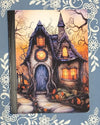 Covered notebook featuring a whimsical illustration of a cozy spooky cottage in an autumn forest on a dark wood desk setup.